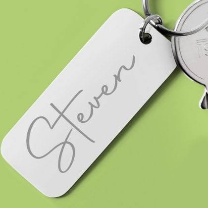 A personalized silver metal key tag engraved with the name 'Steven' in a gray cursive font, attached to a silver keyring against a solid light green background.