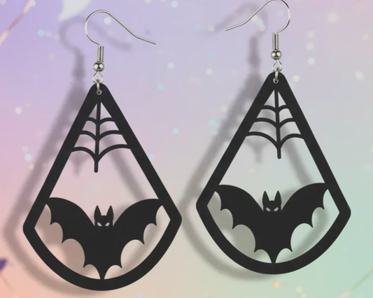 A pair of black teardrop-shaped dangle earrings with laser-cut spiderwebs and bats, hanging from silver fishhook wires against a pastel gradient background with subtle bokeh light effects.