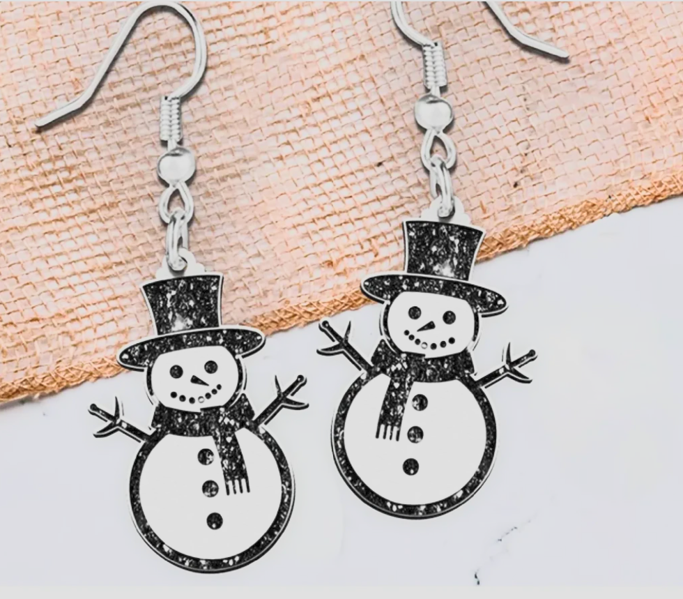 A pair of silver-toned dangle earrings shaped like snowmen, featuring black glittery outlines and details, displayed on a textured beige surface against a white background.