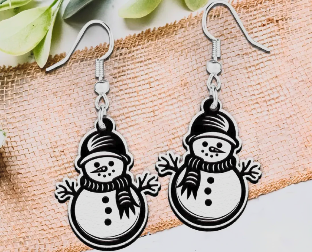 A pair of silver-toned dangle earrings featuring cartoon snowman charms with black outlines and white fill, displayed on a textured beige fabric background with green leaves at the top.