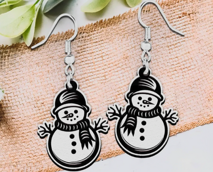 A pair of silver-toned dangle earrings featuring cartoon snowman charms with black outlines and white fill, displayed on a textured beige fabric background with green leaves at the top.